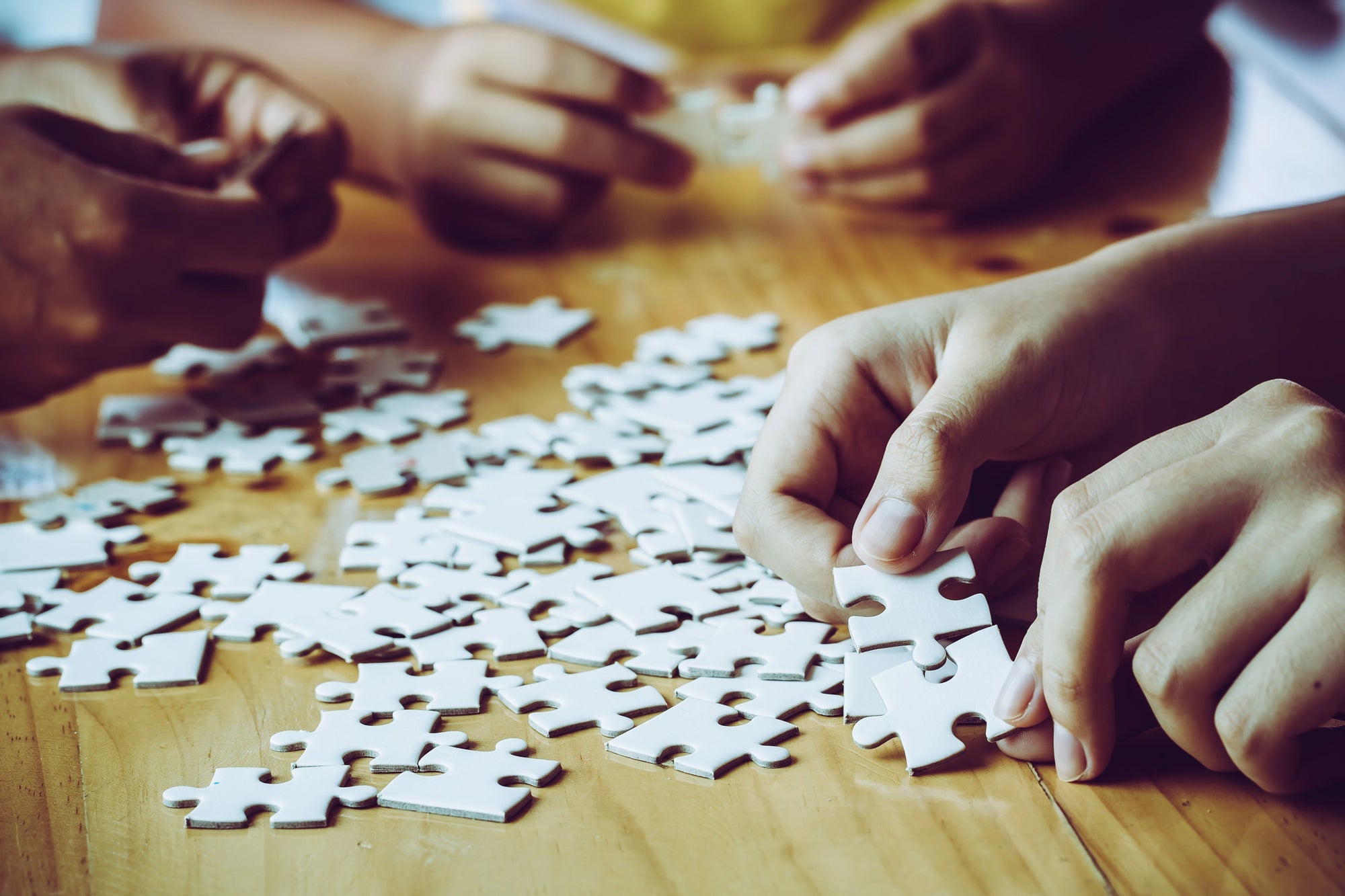 Closeup of the hands of several people all working together on a blank white jigsaw puzzle on a table.