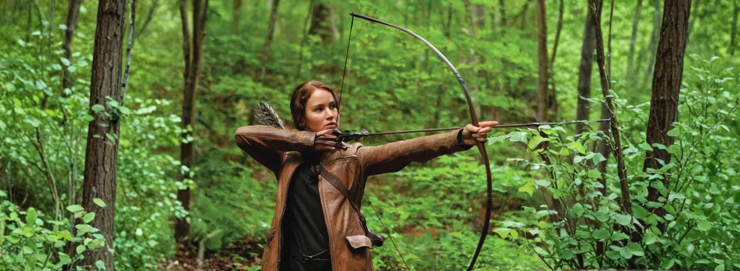 A scene of Katniss Everdeen drawing a bow back while standing in a forest, from the movie Hunger Games.