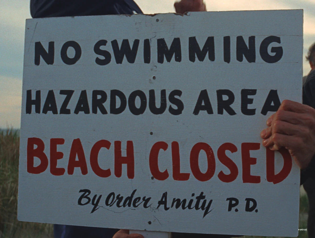 A scene from the film Jaws, showing a closeup of a sign that reads, “No swimming, hazardous area. Beach closed by order Amity P.D.