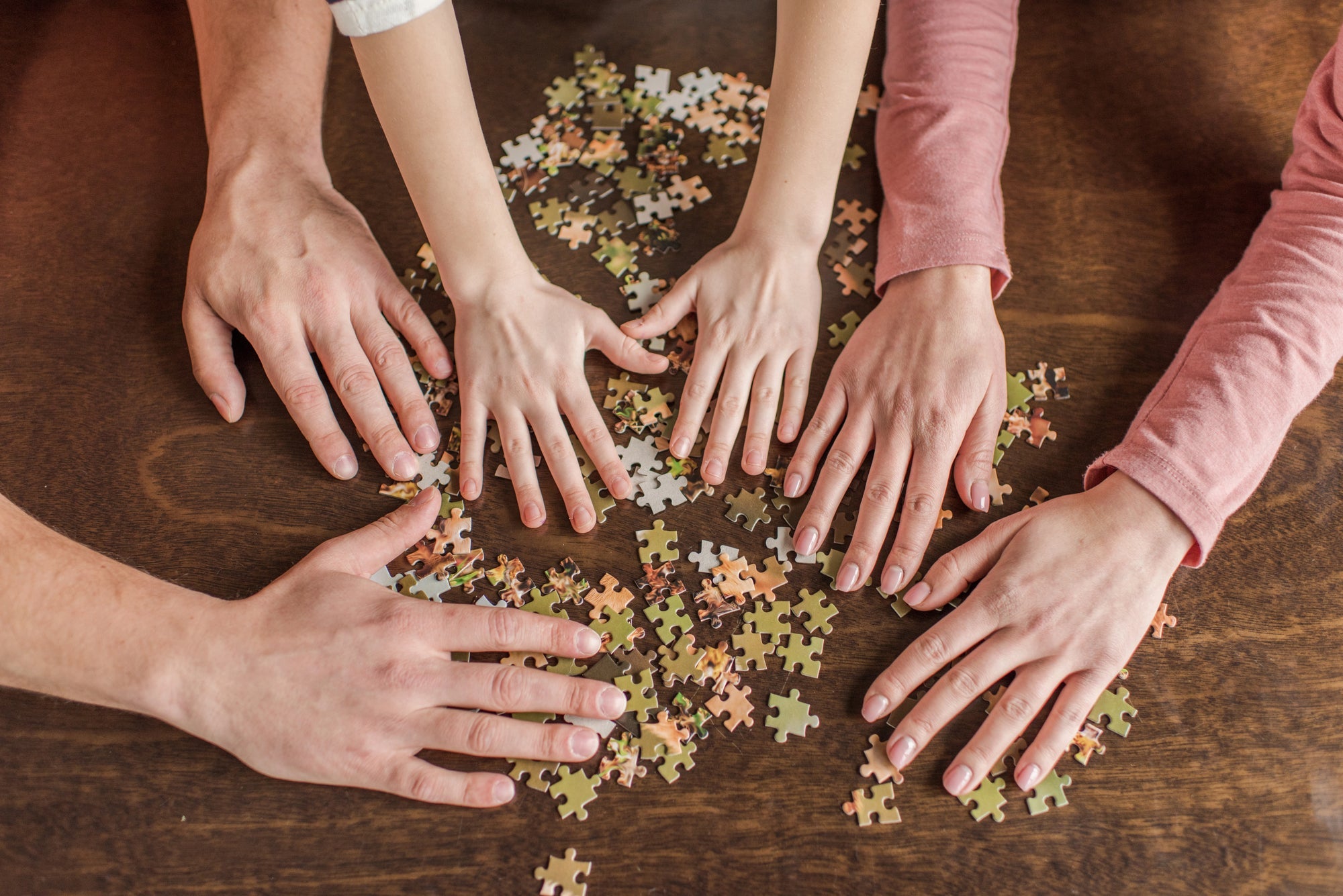 A closeup of a family’s hands as they mix a large pile of jigsaw puzzle pieces together on a table.
