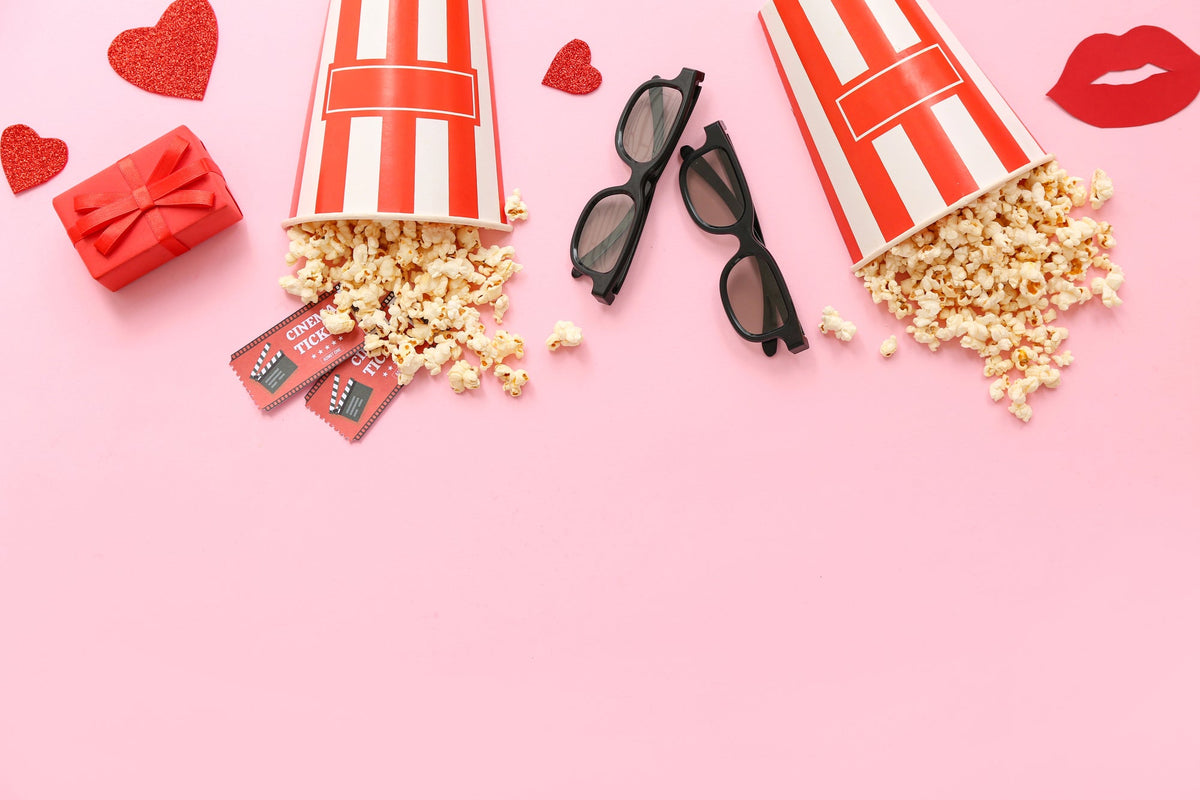 Top-down view of spilled popcorn buckets, 3d glasses, gift boxes, and cinema tickets on a pink background.