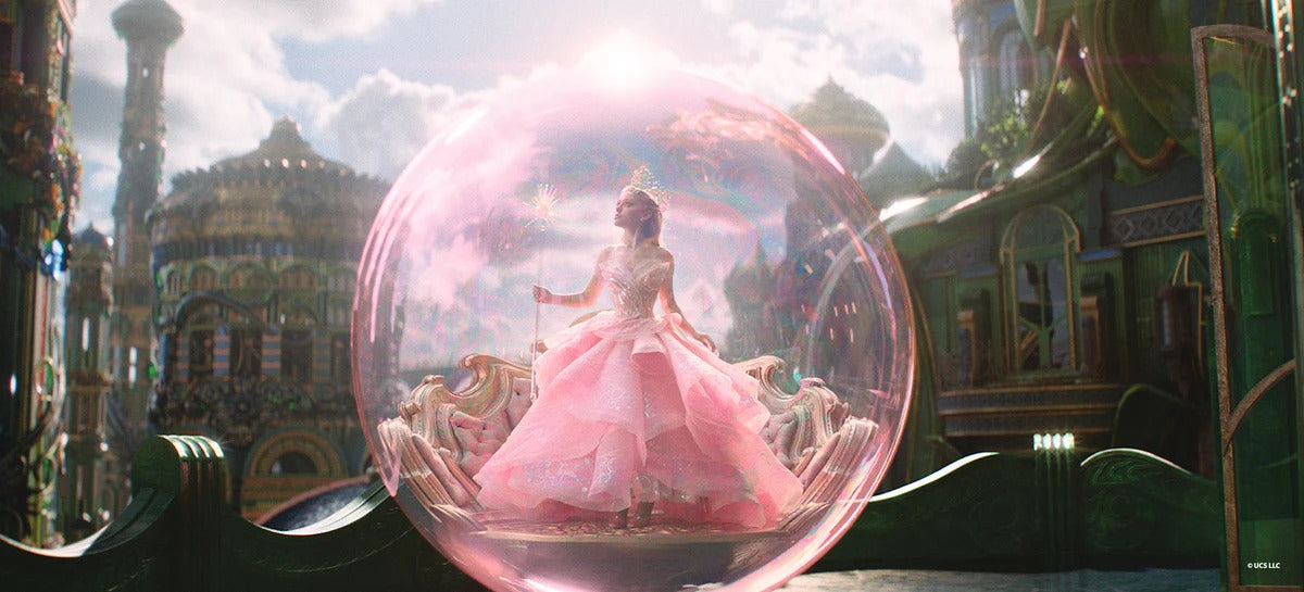 A scene from the film Wicked: For Good, in which Glynda in pink is standing within a shiny pink bubble, surrounded by the greens and golds of the Emerald City landscape.