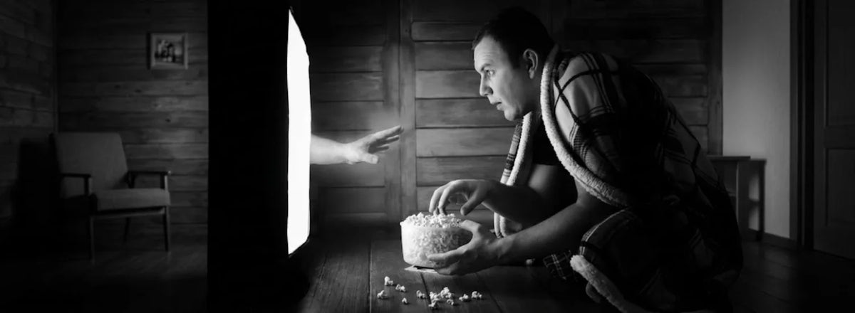 A black and white image of a person watching a tv up close, enraptured, as a spectral hand emerges from the screen to reach toward him.